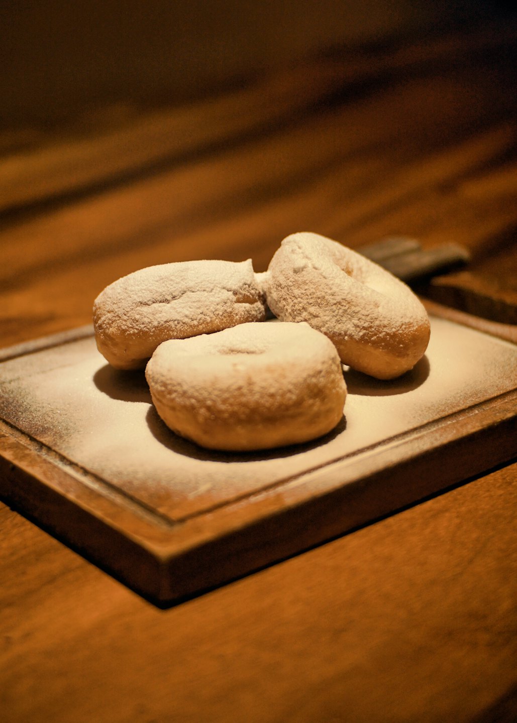 Three powdered sugar donuts sit on a rectangular wooden board.